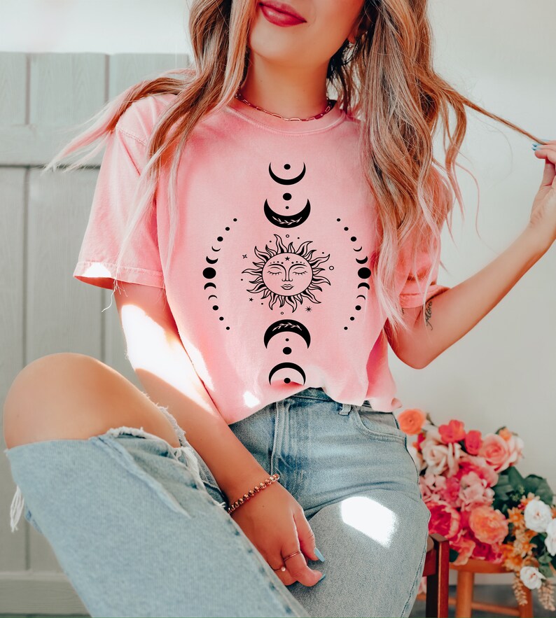 May include: A pink t-shirt featuring a black sun and moon design. The sun has a face and the moon is depicted in various phases.
