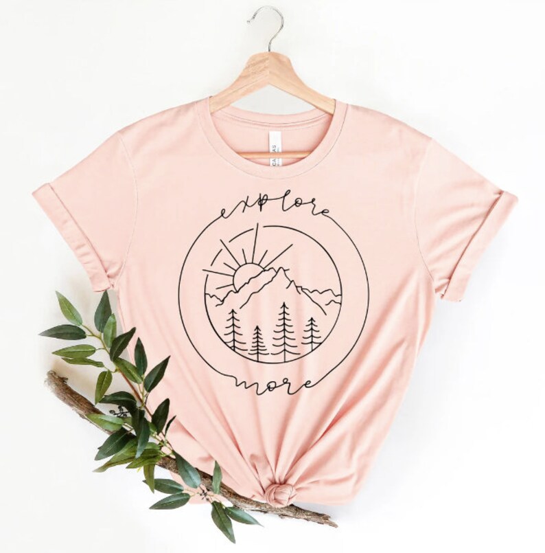 Może przedstawiać: R&oacute;żowy t-shirt z czarnym nadrukiem przedstawiającym pasmo g&oacute;rskie, słońce i drzewa w k&oacute;łku z napisem "explore more" wok&oacute;ł.
