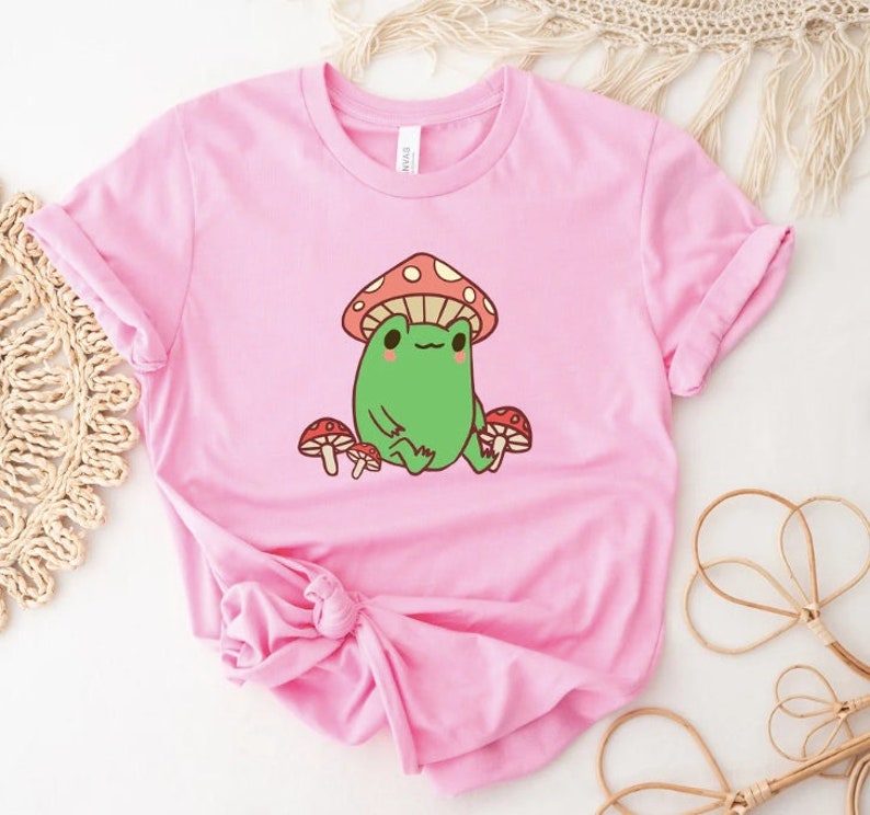 May include: A pink t-shirt with a cartoon frog wearing a red and white mushroom hat. The frog is surrounded by three smaller mushrooms.