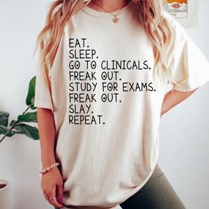 May include: A white t-shirt with black text that reads "EAT. SLEEP. GO TO CLINICALS. FREAK OUT. STUDY FOR EXAMS. FREAK OUT. SLAY. REPEAT."
