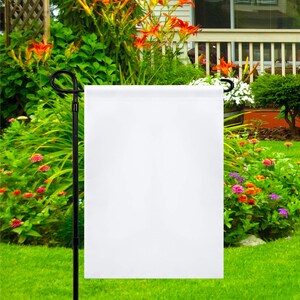 May include: A blank white garden flag is displayed on a black metal flagpole in a green grassy yard. The flag is surrounded by colorful flowers and a white house in the background.