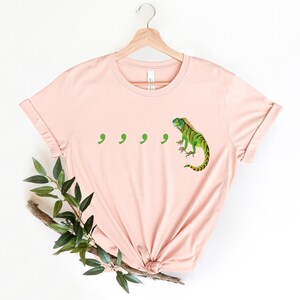 May include: A light pink t-shirt with a green iguana and four green commas printed on the front.
