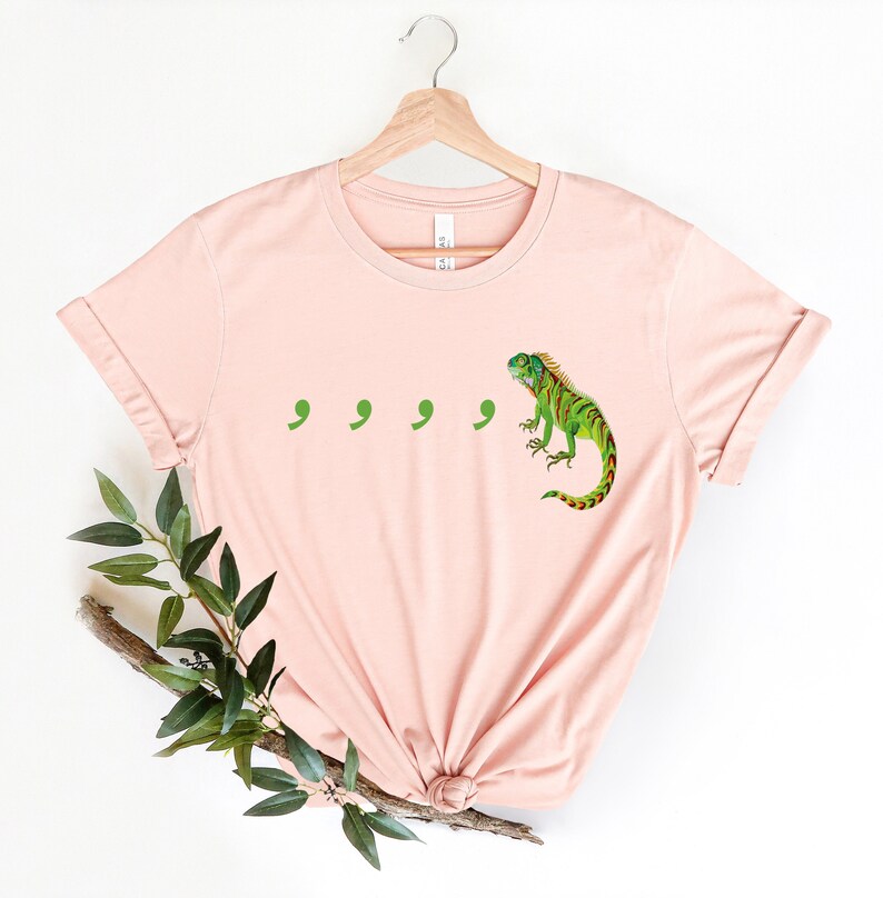 May include: A light pink t-shirt with a green iguana and four green commas printed on the front.