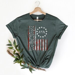 K&ouml;nnte beinhalten: Ein dunkelgr&uuml;nes T-Shirt mit einem abgenutzten Design einer amerikanischen Flagge. Die Flagge ist rot, wei&szlig; und blau mit den Worten "We the People" und dem Jahr "1776" darauf gedruckt.