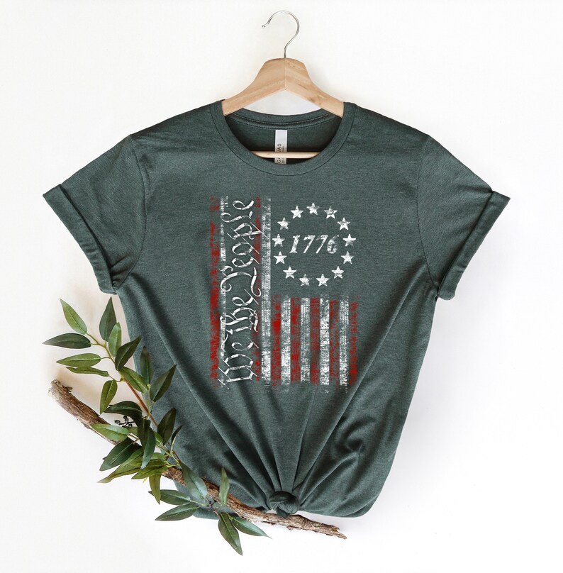 K&ouml;nnte beinhalten: Ein dunkelgr&uuml;nes T-Shirt mit einem abgenutzten Design einer amerikanischen Flagge. Die Flagge ist rot, wei&szlig; und blau mit den Worten "We the People" und dem Jahr "1776" darauf gedruckt.