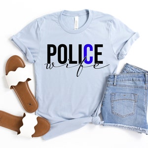 May include: A light blue t-shirt with the text "POLICE wife" in black lettering. The "C" in "POLICE" is blue. The shirt is paired with denim shorts and white sandals.