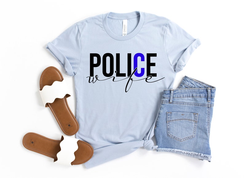 May include: A light blue t-shirt with the text "POLICE wife" in black lettering. The "C" in "POLICE" is blue. The shirt is paired with denim shorts and white sandals.