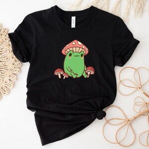 May include: Black t-shirt with a green frog wearing a red and white mushroom hat. The frog is surrounded by three red and white mushrooms.