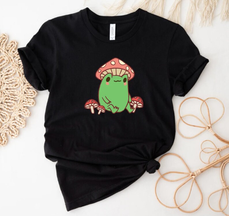 May include: Black t-shirt with a green frog wearing a red and white mushroom hat. The frog is surrounded by three red and white mushrooms.