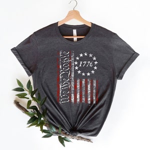 K&ouml;nnte beinhalten: Ein dunkelgraues T-Shirt mit einem abgenutzten Design einer amerikanischen Flagge. Die Flagge zeigt die Worte "We the People" und das Jahr "1776".
