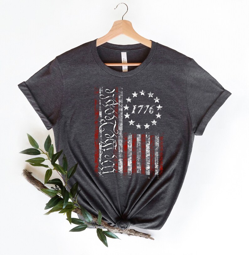 K&ouml;nnte beinhalten: Ein dunkelgraues T-Shirt mit einem abgenutzten Design einer amerikanischen Flagge. Die Flagge zeigt die Worte "We the People" und das Jahr "1776".