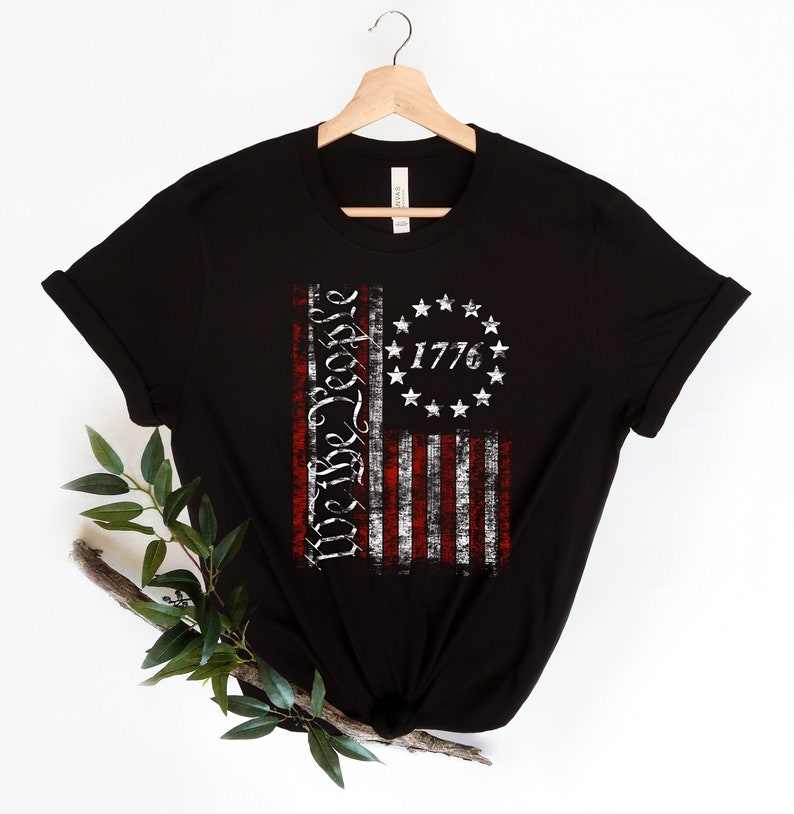 K&ouml;nnte beinhalten: Schwarzes T-Shirt mit einem abgenutzten amerikanischen Flaggen-Design mit den Worten "We the People" und dem Jahr "1776".