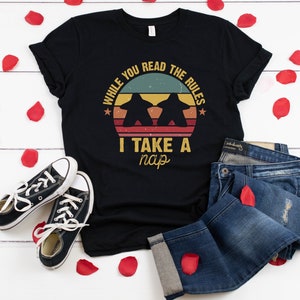 May include: Black t-shirt with a vintage-style graphic that says "While you read the rules I take a nap". The graphic features two game pieces and a sunset.