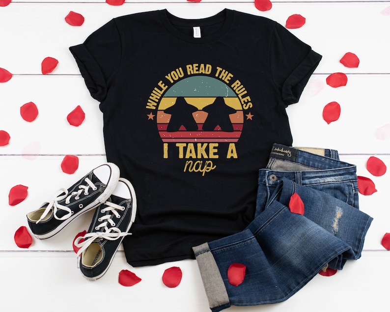 May include: Black t-shirt with a vintage-style graphic that says "While you read the rules I take a nap". The graphic features two game pieces and a sunset.