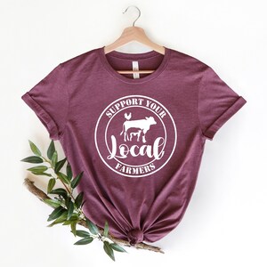May include: A maroon t-shirt with a white circle graphic that says "Support Your Local Farmers" with a cow, calf, pig, and chicken inside the circle.