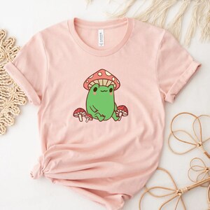 May include: A pink t-shirt with a cartoon frog wearing a red and white mushroom hat. The frog is surrounded by three red and white mushrooms.