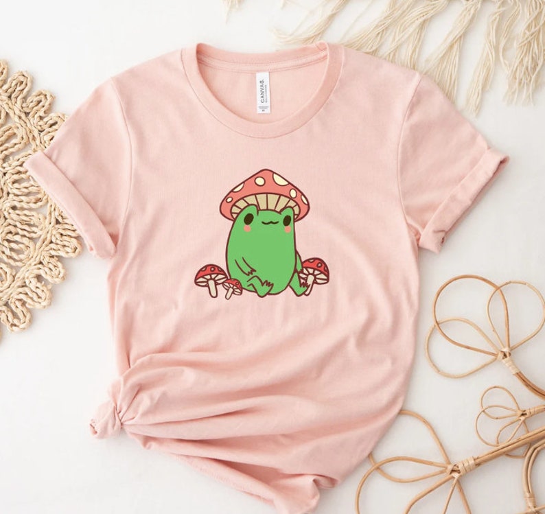 May include: A pink t-shirt with a cartoon frog wearing a red and white mushroom hat. The frog is surrounded by three red and white mushrooms.