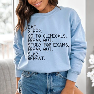 May include: Light blue sweatshirt with the text "EAT. SLEEP. GO TO CLINICALS. FREAK OUT. STUDY FOR EXAMS. FREAK OUT. SLAY. REPEAT."