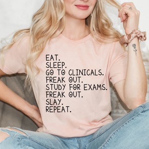 May include: A pink t-shirt with black text that reads "EAT. SLEEP. GO TO CLINICALS. FREAK OUT. STUDY FOR EXAMS. FREAK OUT. SLAY. REPEAT."
