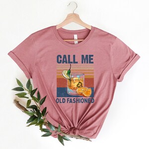 May include: A pink t-shirt with the text "CALL ME OLD FASHIONED" and a vintage-style illustration of an old fashioned cocktail with a lemon wedge, orange slices, and a green chili pepper.