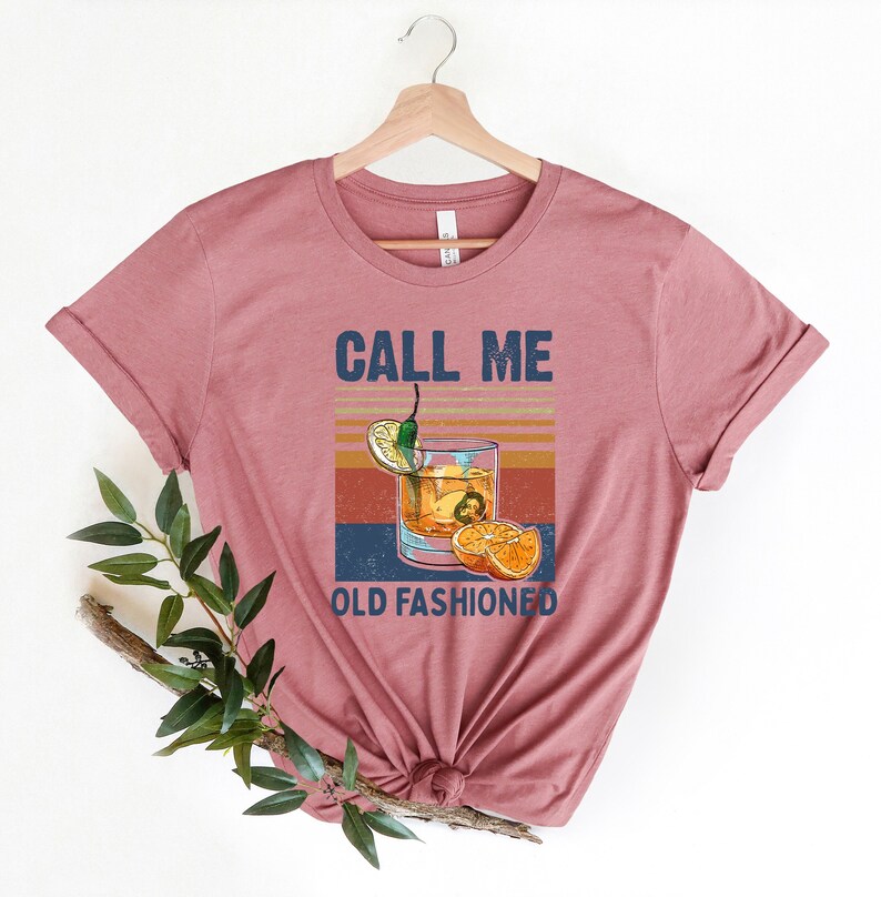 May include: A pink t-shirt with the text "CALL ME OLD FASHIONED" and a vintage-style illustration of an old fashioned cocktail with a lemon wedge, orange slices, and a green chili pepper.