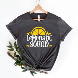 May include: A dark gray t-shirt with a white graphic that says "Lemonade Squad" and a yellow lemon slice graphic.