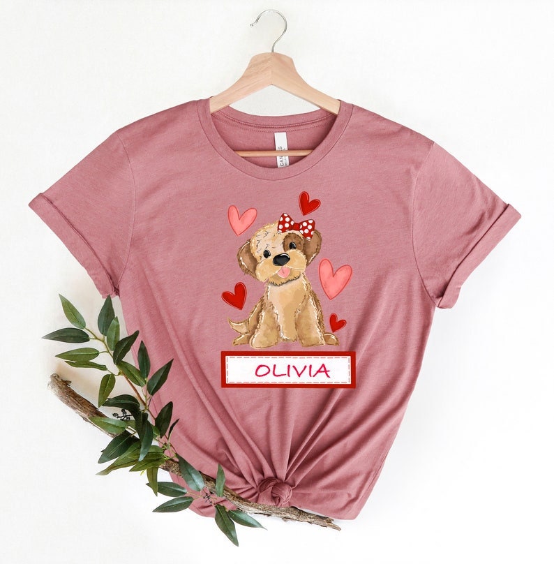 K&ouml;nnte beinhalten: Ein pinkfarbenes T-Shirt mit einer Cartoon-Illustration eines braunen Hundes, der eine rote Schleife mit wei&szlig;en Punkten tr&auml;gt. Der Hund ist von roten Herzen umgeben. Der Text "OLIVIA" ist auf einem roten Banner unter dem Hund gedruckt.