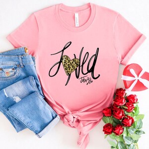 May include: Pink t-shirt with a black and gold leopard print heart and the word "Loved" written in black script. The text "John 3:16" is written in black script below the heart.