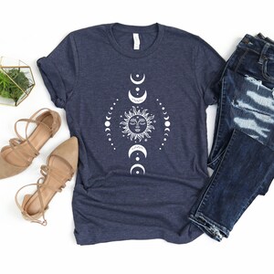 May include: A navy blue t-shirt featuring a white sun and moon design. The design depicts a sun with a face and a circle of moons surrounding it. The t-shirt is laid flat on a white background with a pair of brown sandals and blue jeans.