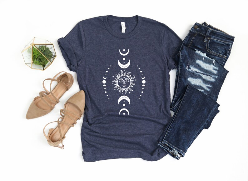 May include: A navy blue t-shirt featuring a white sun and moon design. The design depicts a sun with a face and a circle of moons surrounding it. The t-shirt is laid flat on a white background with a pair of brown sandals and blue jeans.