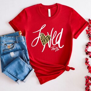 May include: Red short-sleeved t-shirt with a white graphic that says "Loved" with a leopard print heart and the text "John 3:16".