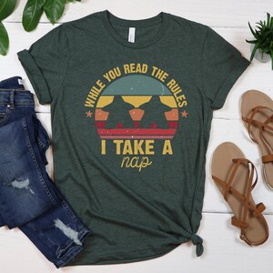 May include: A green t-shirt with a vintage-style graphic that says "While you read the rules I take a nap". The graphic features a sunset with two silhouettes of dogs.