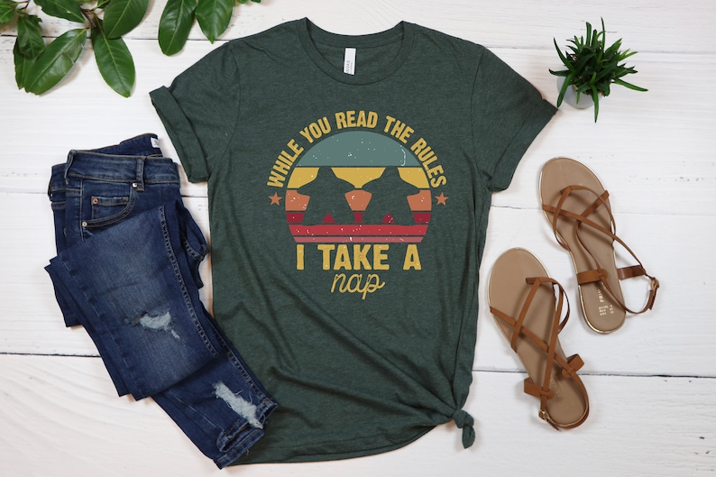 May include: A green t-shirt with a vintage-style graphic that says "While you read the rules I take a nap". The graphic features a sunset with two silhouettes of dogs.