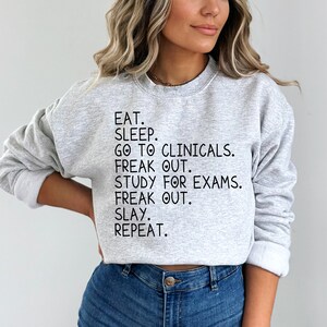 May include: A gray sweatshirt with the text "EAT. SLEEP. GO TO CLINICALS. FREAK OUT. STUDY FOR EXAMS. FREAK OUT. SLAY. REPEAT." printed in black.