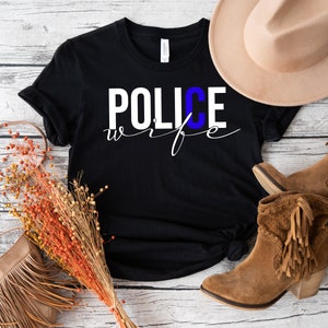 May include: Black t-shirt with white text that reads "POLICE wife" with a blue "C". The shirt is laid out on a white wooden surface with a brown hat, a brown purse, and brown boots.