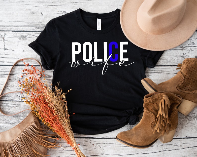 May include: Black t-shirt with white text that reads "POLICE wife" with a blue "C". The shirt is laid out on a white wooden surface with a brown hat, a brown purse, and brown boots.