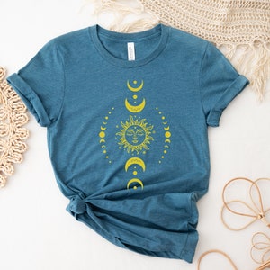 May include: A teal blue t-shirt featuring a yellow sun and moon design. The design depicts a sun with a face and a circle of moons surrounding it.