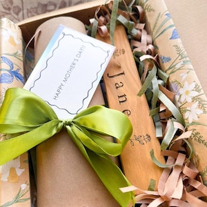 May include: A gift box with a green satin bow, containing a card that reads "HAPPY MOTHER'S DAY!!" and a wooden spatula engraved with the name "Jane". The box is filled with decorative paper and ribbons.