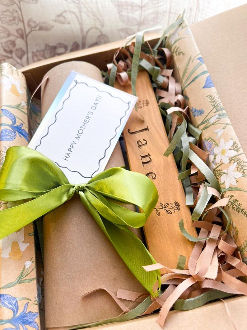May include: A gift box with a green satin bow, containing a card that reads "HAPPY MOTHER'S DAY!!" and a wooden spatula engraved with the name "Jane". The box is filled with decorative paper and ribbons.