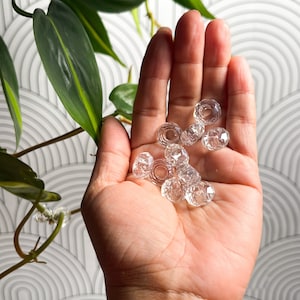 May include: A hand holding a collection of clear glass beads with faceted surfaces. The beads are round and have a hole in the center.