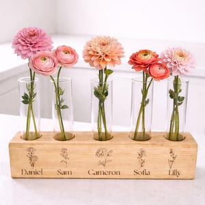 May include: A wooden flower vase with five glass tubes, each holding a different colored flower. The wooden base is engraved with names: Daniel, Sam, Cameron, Sofia, and Lilly. The flowers are pink, peach, and orange.