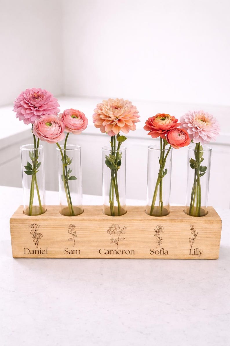 May include: A wooden flower vase with five glass tubes, each holding a different colored flower. The wooden base is engraved with names: Daniel, Sam, Cameron, Sofia, and Lilly. The flowers are pink, peach, and orange.