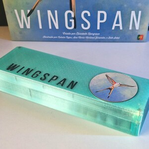 May include: A rectangular, translucent light-blue storage box featuring the word WINGSPAN printed in black letters on the top surface. A circular sticker depicting a bird in flight is attached to the right side of the lid, positioned below the game box.