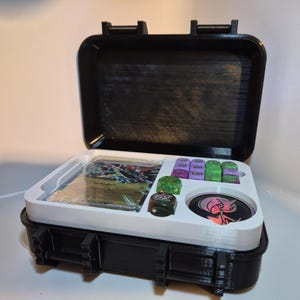 Pode incluir: Uma mala de transporte preta de revestimento rígido com um inserto interior branco contendo uma carta colecionável numa manga protetora, seis dados pequenos roxos e verdes, um dado verde brilhante, um dado escuro com um símbolo EX e uma ficha de jogo circular.