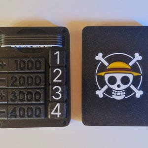 Puede incluir: Un contador de juego rectangular negro con un teclado numérico que muestra signos de más y menos junto a los valores 1000, 2000, 3000 y 4000. Al lado, una cubierta rectangular negra muestra un logotipo de calavera blanca con un sombrero de paja amarillo.