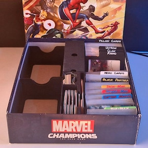 May include: A Marvel Champions The Card Game box with a black internal organizer. The box features labeled dividers for Villain Cards, Hero Cards, Black Panther, Iron-Man, Spider-Man, Captain Marvel, and She-Hulk, with empty compartments.