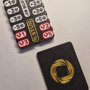 May include: A black rectangular game tracker with white and red inserts featuring numerical values and symbols, including text labels like BUFF and STUN. Beside it lies a black rectangular card with a gold circular emblem in the center.