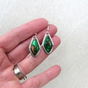 May include: A pair of dangle earrings featuring a diamond-shaped green stone with gold veining, set in a silver-colored frame. The earrings have a decorative, textured border and a simple hook closure.