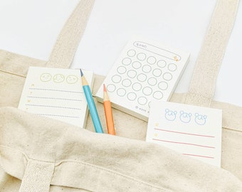 May include: A beige canvas tote bag with three different notepads inside. The notepads have different designs, including a checklist, a weekly planner, and a notepad with cute cartoon faces. Two colored pencils are also inside the bag.