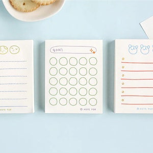 May include: Three white notepads with different designs. The first notepad has a weekly planner with a blue and gold design. The second notepad has a goal tracker with a green and orange design. The third notepad has a lined notepad with a red and gold design.